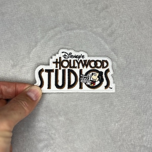 May include: A Disney's Hollywood Studios patch with a white border. The patch features the text "Disney's Hollywood Studios" in brown and black, with Mickey Mouse holding a film clapper. The patch is held by a hand.