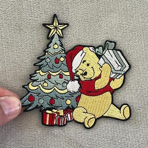 Peut inclure: Écusson brodé représentant un ours jaune portant un bonnet de Père Noël et une écharpe rouge, portant un cadeau emballé. Un sapin de Noël décoré et des cadeaux sont également dans le design. Le patch a une bordure noire.
