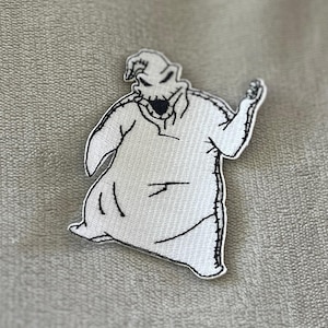 May include: A white embroidered patch featuring the character Oogie Boogie from The Nightmare Before Christmas. The patch has a black outline and stitching, depicting the character's form and facial features. The patch is on a textured, light gray surface.