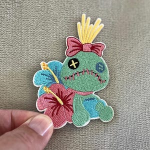 May include: Embroidered patch of a cartoon character in green and blue, with a pink bow and hibiscus flower. The patch has a stitched design and a yellow crown. The character's face has stitched details.