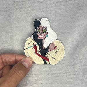 Puede incluir: Un parche bordado que muestra un personaje con cabello dividido en negro y blanco, sombra de ojos verde y una sonrisa roja. El personaje viste un abrigo de piel color crema y guantes rojos, sosteniendo una boquilla larga y azul claro.