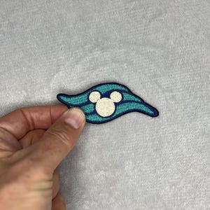 以下が含まれることがあります： 青と白の刺繍パッチ。ミッキーマウスの頭部が描かれています。パッチは波型のデザインで、水平ストライプと濃い青色の縁取りがあります。ミッキーマウスの頭部は白です。