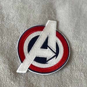 Może przedstawiać: Biała naszywka haftowana z czerwonym i niebieskim kółkiem wokół litery "A".