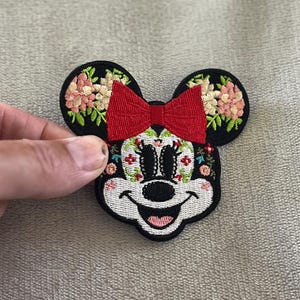 May include: Embroidered patch featuring a stylized mouse face with black ears and a red bow. The mouse face is decorated with floral patterns and a smiling expression. The patch is likely for clothing or accessories.