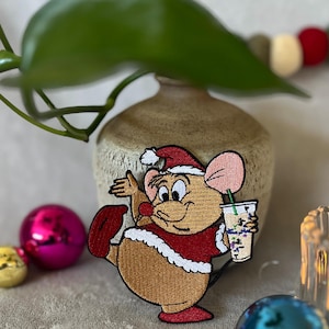 以下が含まれることがあります： サンタ帽と赤い衣装を着た漫画のネズミが飲み物を持っている刺繍パッチ。 ネズミは茶色で、ピンクの耳と赤い鼻をしています。 パッチは白い縁取りがあり、装飾的なオーナメントが付いたニュートラルな背景に配置されています。