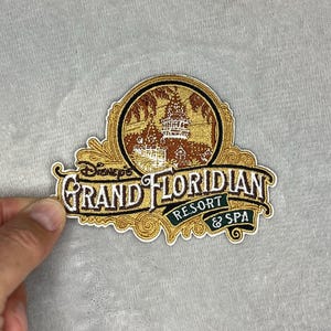 Könnte beinhalten: Gestickter Aufnäher mit der Aufschrift "Disney's Grand Floridian Resort & Spa". Der Aufnäher hat einen goldenen Rand und ein rundes Bild eines Gebäudes mit Palmen. Der Text ist in Gold und Weiß gehalten.