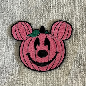Könnte beinhalten: Ein rosa-schwarzer, gestickter Aufnäher in Form eines Kürbisses mit Mickey Mouse Ohren. Der Kürbis hat ein lächelndes Gesicht und grüne Blattakzente. Geeignet für Kleidung oder Accessoires.