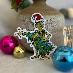 May include: Embroidered patch featuring a green Grinch character wearing a Santa hat and holding a Christmas tree. The patch has a white border and is surrounded by Christmas ornaments in pink, gold, and blue.