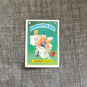 Pode incluir: Um cartão colecionável Garbage Pail Kids com um bebê de desenho animado chamado Jason Basin sentado em um vaso sanitário. O cartão tem um fundo verde e o texto "Garbage Pail Kids" no topo.
