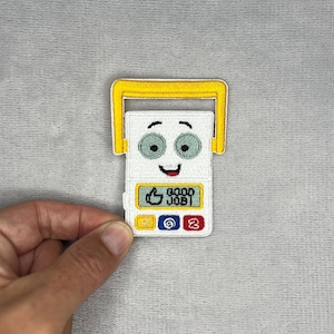 May include: An embroidered patch shaped like a white rectangular device with a yellow handle. It features a cartoon face with green eyes and a smiling mouth. Below the face is a yellow box with a thumbs-up icon and the text GOOD JOB, plus three small coloured buttons.