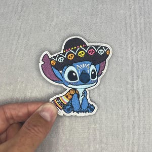 May include: An embroidered patch featuring a blue alien character wearing a black sombrero decorated with colorful skull patterns. The character has large dark eyes, purple inner ears, and a striped serape draped over one shoulder with a white border.