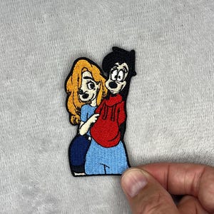 May include: Embroidered patch featuring two cartoon characters, a blonde woman in a blue dress and a man in a red hoodie. The patch has a black border and is likely for clothing or accessories.