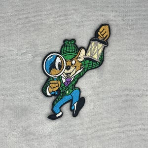 May include: An embroidered patch featuring a cartoon mouse dressed as a detective in a green plaid hat and coat, a purple tie, and blue trousers. The mouse holds a magnifying glass in one hand and a raised lantern in the other, outlined in black.