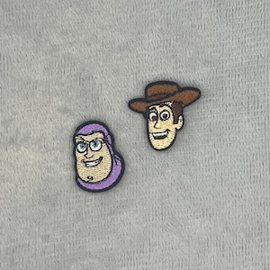 Puede incluir: Dos parches de tela bordados con los rostros de dos personajes animados. Un parche muestra a un personaje con capucha morada y ojos azules, mientras que el otro muestra a un personaje con un sombrero de vaquero marrón y cara bronceada.