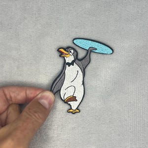May include: An embroidered patch featuring a cartoon penguin with a white belly, grey back, and a black bowtie. The penguin is standing on one foot, holding a light blue oval-shaped tray or platter above its head with one raised wing.