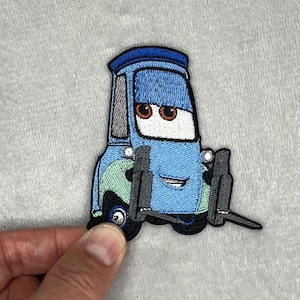May include: Embroidered patch featuring a cartoon car character in shades of blue, with black wheels and a smiling face. The patch has a detailed design, including the car's headlights and a tow hook.
