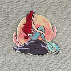 May include: Embroidered patch featuring a mermaid with red hair, a turquoise tail, and a light blue top. The mermaid is seated on a dark gray rock, with a peach-colored background and pink coral accents. The patch has a white border.