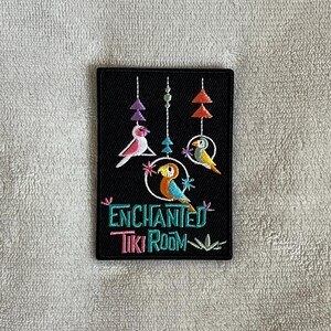 May include: Black embroidered patch with a tiki room sign. The sign says "Enchanted Tiki Room" with a colorful bird design above the text.