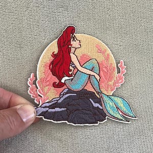 May include: Embroidered patch featuring a mermaid with red hair, a blue tail, and a seashell top, sitting on a dark gray rock. The mermaid is set against a light orange background with pink coral accents.