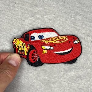 May include: Embroidered patch featuring a red race car with the number 95 on the side. The car has blue eyes, a smiling mouth, and the words "Rust-eze" on the hood. The patch is likely for clothing or accessories.