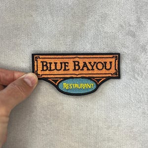 Puede incluir: Parche bordado con las palabras "BLUE BAYOU" en naranja, sobre la palabra "RESTAURANT" en un óvalo azul. El parche tiene un borde negro y está sobre un fondo gris.