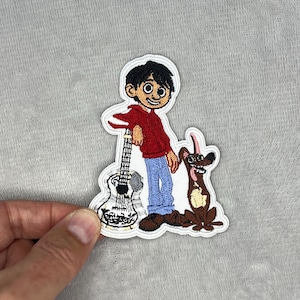 May include: Embroidered patch featuring a cartoon character in a red jacket and blue pants, leaning on a guitar. A brown dog with a pink tongue sits beside him. The patch has a white border and is held by a hand.