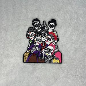 May include: Embroidered patch featuring a family of skeletons in vibrant colors. The patch depicts a group of skeletons with detailed facial features and clothing, with a Day of the Dead theme. The patch is on a light gray background.