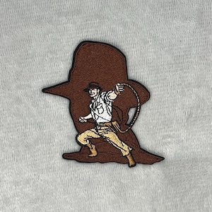 May include: Embroidered patch featuring a figure in a hat and whip, set against a brown silhouette. The figure is wearing a white shirt, tan pants, and brown boots. The patch is likely for clothing or accessories.