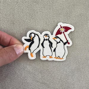 May include: Embroidered patch featuring three penguins. The penguins are black and white with orange feet and beaks. One penguin is dancing, another is standing, and the third is holding a red umbrella with a white floral design.