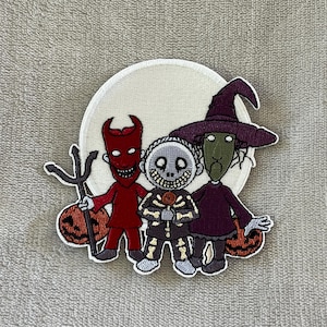 May include: Embroidered patch featuring three characters from a Halloween-themed movie. The characters include a red devil with a trident, a skeleton, and a witch. The patch is set against a white circle background with two orange pumpkins.
