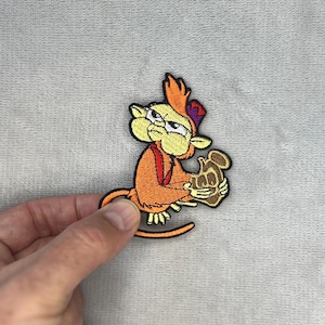 Pode incluir: Patch bordado com um macaco de desenho animado em tons de laranja, amarelo e castanho. O macaco usa um chapéu roxo e segura um objeto castanho. O patch tem uma borda preta. O fundo é cinzento.