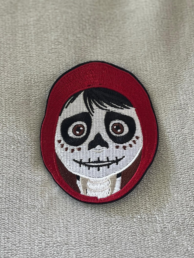 Miguel patch from Coco with painted skull face & red hoodie. 3.5x2.75 inch embroidered iron-on — perfect Disney Día de los Muertos accent.