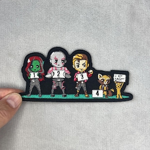 以下が含まれることがあります： 人気のSF映画のキャラクターが描かれた刺繍パッチ。パッチには4人のキャラクターがおり、それぞれに番号が振られており、1人は「I AM GROOT」と書かれた看板を持っています。キャラクターは黒い背景に並んでいます。
