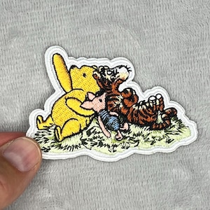 May include: Embroidered patch featuring Winnie the Pooh, Piglet, and Tigger. Pooh is yellow, Piglet is pink, and Tigger is orange and brown. The patch is on a white background with green grass.