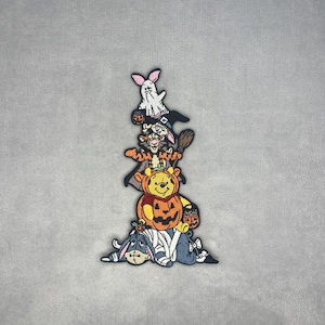 May include: An embroidered patch featuring a vertical stack of cartoon characters dressed in Halloween costumes. At the base is a blue donkey wrapped in bandages, topped by a yellow bear inside a pumpkin, an orange tiger with a broom, and a white ghost.
