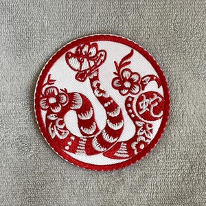 Peut inclure: Patch brodé rouge avec un motif de serpent blanc. Le serpent est enroulé autour d'une fleur et présente un motif rayé rouge et blanc.
