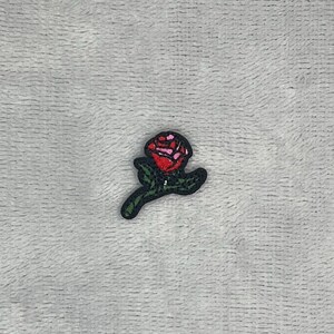 May include: A small embroidered patch featuring a stylised rose with a vibrant red and pink flower head and a dark green stem with leaves. The design is outlined in black thread, creating a clear and defined shape against the textured background.