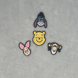May include: Four embroidered fabric patches featuring the faces of cartoon characters. The set includes a blue donkey, a yellow bear, a pink piglet, and an orange and black striped tiger, all outlined in black thread on a textured gray background.