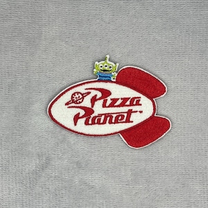 May include: An embroidered patch shaped like a rocket ship with a white background and red border. The text Pizza Planet is stitched in red with a small planet icon. A small green three-eyed alien character is positioned on top of the rocket.