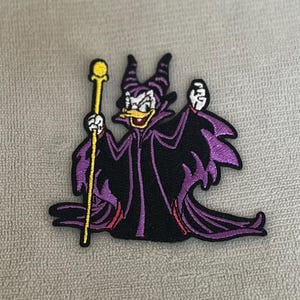 Könnte beinhalten: Lila und schwarzer gestickter Bügelaufkleber mit einer Cartoonfigur mit einem gelben Stab. Die Figur trägt eine lila Robe, Hörner und einen schelmischen Ausdruck.