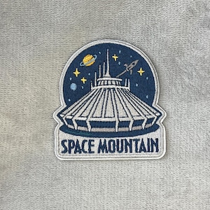 May include: Embroidered patch featuring the Space Mountain attraction. The patch has a dark blue background with a white and gray Space Mountain design, a yellow planet, stars, and a rocket. The text "SPACE MOUNTAIN" is displayed in white.