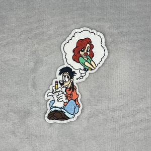 May include: An embroidered patch featuring a cartoon character with black hair wearing an orange hoodie and blue pants, holding a yellow pencil and paper. Above the character is a white thought bubble containing an illustration of a red-haired character.