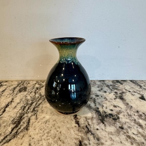 May include: A handcrafted ceramic vase with a dark blue glaze. The vase has a rounded body and a flared neck with a gradient of green and brown. The base is a lighter brown color. The vase is approximately 7 inches tall.