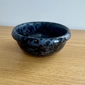 Pode incluir: Uma tigela pequena e redonda com um padrão azul e preto salpicado. A tigela tem uma borda lisa e arredondada e um interior escuro. A tigela está sobre uma superfície de madeira castanha clara.