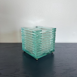 May include: A square-shaped decorative container constructed from stacked layers of clear, light green glass. The structure features a ribbed, horizontal design and is supported by four small, metallic, spherical feet at the base corners.