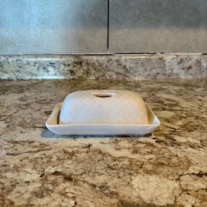 May include: A white ceramic butter dish with a lid featuring a diamond pattern. The dish has a rectangular base with rounded corners and small handles. The lid has a small opening and a matching diamond pattern.