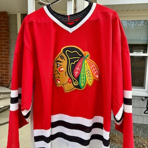 Puede incluir: Camiseta de hockey roja con rayas blancas y negras. La camiseta presenta un gran logotipo bordado de una cabeza de nativo americano con un tocado de plumas. El logotipo es de color rojo, negro, verde y amarillo.