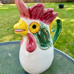 Puede incluir: Una jarra de cerámica blanca con forma de gallo con detalles pintados en rojo, amarillo y verde. El gallo tiene un pico grande y un ojo amarillo brillante.