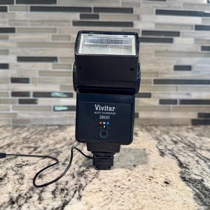 以下が含まれることがあります： 白いフラッシュヘッドと黒いコードが付いた、黒いVivitar Auto Thyristor 2800フラッシュユニットです。