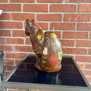 May include: A brown ceramic cat figurine with painted flowers, hearts, and vines. The cat is sitting on a black glass table with a silver metal frame.